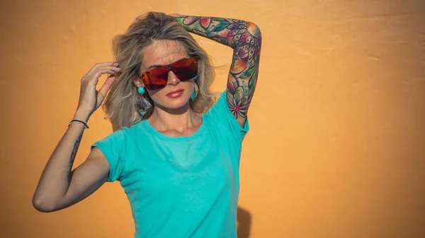 Gyönyörű szőke napszemüveg pózol a kamera. Portré a háttéren világos narancssárga fal. Modern csípő lány tetoválás. Vértes portré. — Stock Fotó