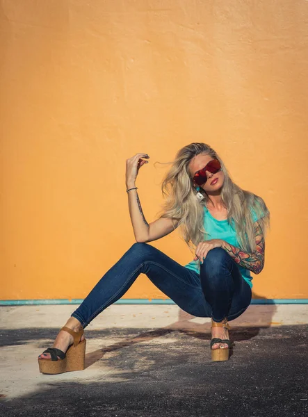 Vacker blondin i solglasögon poserar på kameran. Porträtt på bakgrunden av ljusa orange vägg. Moderna hipster flicka med tatueringar. Sittande porträtt — Stockfoto