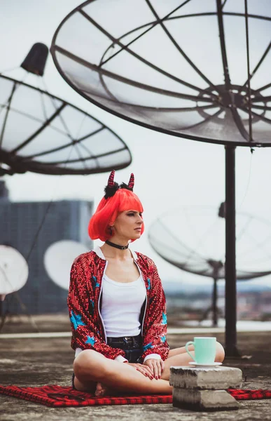 Gyönyörű lány az ördög ül a tető a ház Halloween számítva. Portré a háttérben a nagy műholdas antennák. Modern divat — Stock Fotó