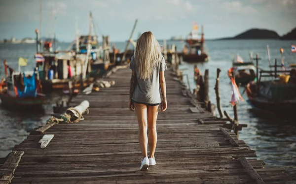 Met lang blond haar komt door de oude houten pier in de stralen van de ondergaande zon. Terug naar de camera. Vakantie in Azië. — Stockfoto