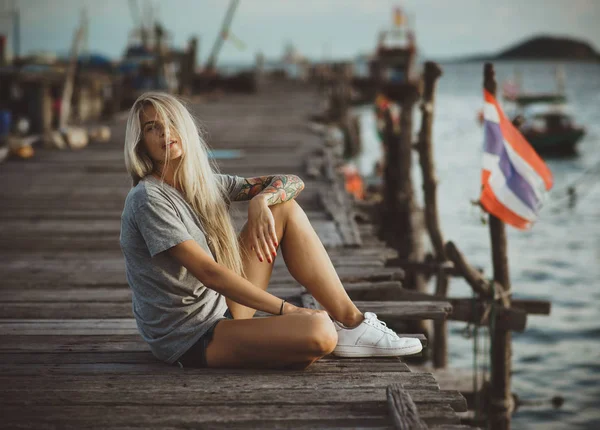 Hipster menina sentada no cais de madeira nos raios do sol poente. Umas férias na Tailândia. Mãos com tatuagens — Fotografia de Stock