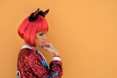 Güzel kız Halloween beklentisiyle duvara karşı duran kırmızı şeytan kostümlü. Kırmızı peruk ve boynuzları takar. Renk kavramı