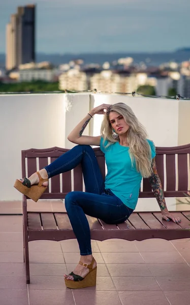 Linda loira posando para a câmera sentada em um banco de madeira no telhado. Usa jeans e sapatos em uma plataforma alta. O conceito de cor turquesa. Mulher moderna . — Fotografia de Stock