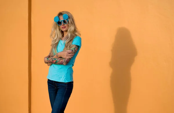 Vacker blondin i solglasögon poserar på kameran. Porträtt på bakgrunden av ljusa orange vägg. Moderna hipster flicka med tatueringar. — Stockfoto