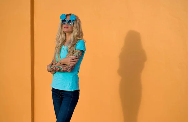 Mooie blonde in zonnebril poseren op de camera. Portret op de achtergrond van de heldere oranje muur. Moderne hipster meisje met tatoeages. — Stockfoto