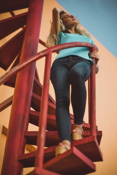 Красивая длинноногая блондинка стоит на красной спиральной лестнице и смотрит вдаль. На фоне оранжевых стен и неба. Современная мода — стоковое фото