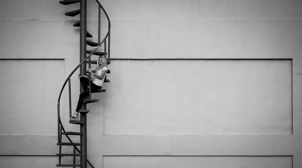 Красивая длинноногая блондинка сидит на красной спиральной лестнице и смотрит вдаль. На фоне оранжевых стен и неба. Современная уличная мода Черное и белое — стоковое фото