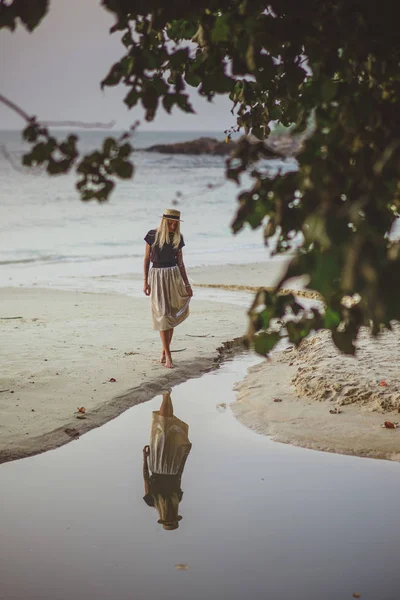 En ung kvinna står vid lugnvattnet tidigt på morgonen. Sandstrand med lummiga gröna träd. En semester i Thailand. — Stockfoto