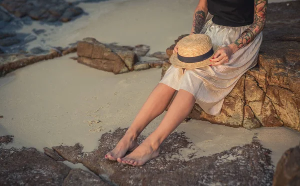 Lány ül a kövek reggel tenger partján gazdaság térdre egy szalmakalapot. Hosszú világos szoknyát visel. Reggel sétál a tengerparton — Stock Fotó