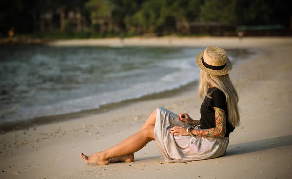 Ung kvinna sitter på morgonen av sandstranden. Bär en stråhatt, lång kjol och svart t-shirt. morgonpromenad — Stockfoto