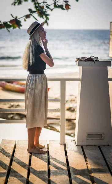 Blondin i lång kjol stående på träbron på bakgrunden av havets morgon och justerar hennes örhängen. En morgonpromenad vid havet — Stockfoto