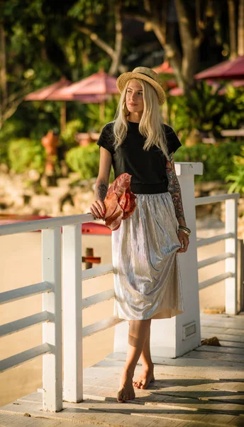 Vackra blonda promenader på morgonen stranden med gula blad i hennes händer. Bär en stråhatt, lång kjol och svart t-shirt. Semester i Thailand — Stockfoto