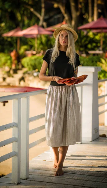 Beautiful blonde walking on the morning beach with yellow leaves in her hands. Wears a straw hat, long skirt and black t-shirt. Vacation in Thailand — Stock Photo, Image