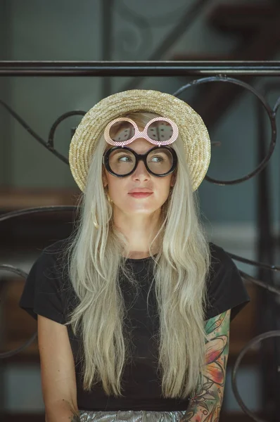 Loira bonita com um sorriso encantador, sentada na sala de estar brilhante com óculos redondos. Usa um chapéu de palha e t-shirt preta — Fotografia de Stock