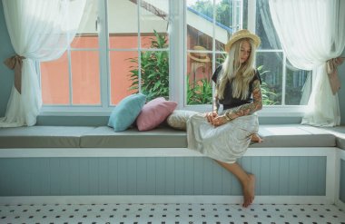 Sarışın Hasır Şapkalı bir pencere kenarındaki kanepede oturuyor. modern bir kız görüntüsü. Dövmeler ile model