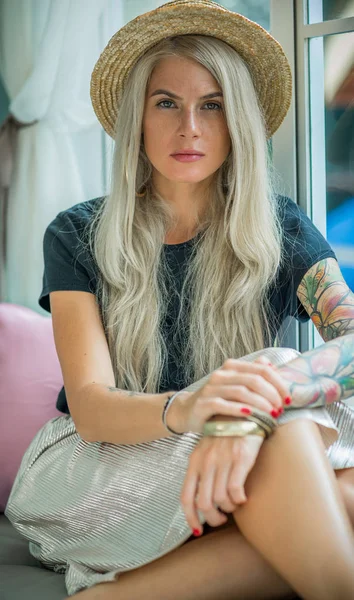 Blonde in a straw hat sits on the couch by the window. the image of a modern girl. Model with tattoos. closeup — Stock Photo, Image