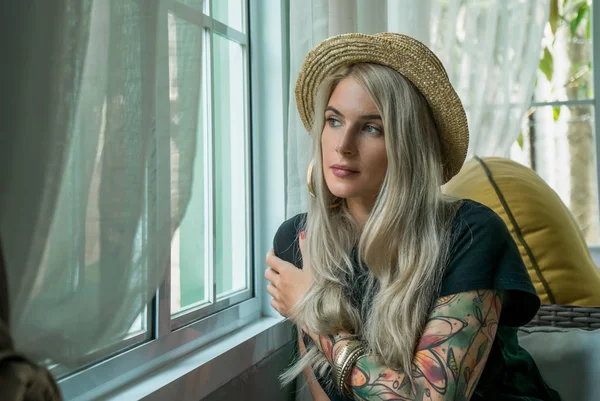 Beautiful blonde with dreamy views sits at the window and looks outside. image of the modern girl. Tattoo model. Closeup portrait — Stock Photo, Image