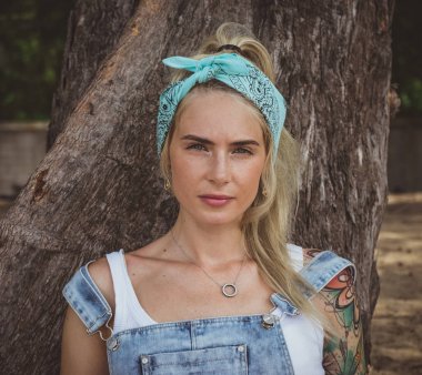 Güzel model bir ağaç yakınındaki plajda otururken dövmeli. Turkuaz bir bandana giyer. Modern bir kadın. Cennette huzur.