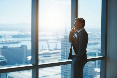 Handsome mature business executive standing at large windows in a top floor office, talking on his mobile phone while looking out the view of a city below
