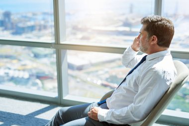 Mature businessman sitting comfortably in his office, talking on his mobile phone while looking out of his windows at the city below clipart