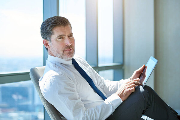 Portrait of a mature CEO sitting in his office, comfortably using a digital tablet, and looking at the camera with a serious yet positive expression