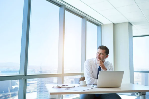 Handsome mature business leader sitting in a modern office with large windows, at his tidy desk with laptop open, looking out through the window with a positive expression and gentle sunflare