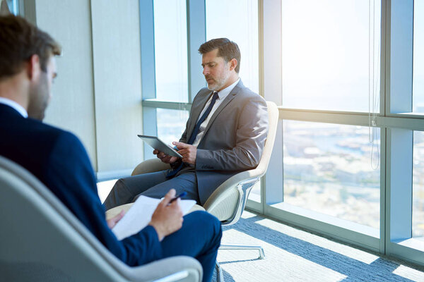 Mature business executive reading information on a digital tablet, with younger businessman filling out information on a form