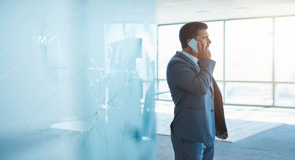 Mature businessman in a new modern office talking on his mobile phone while wearing a corporate business suit and looking away at large windows