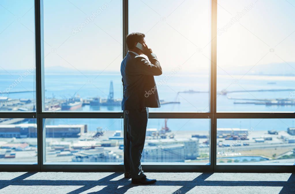 Mature business executive standing next to large windows in a top floor office looking at the view of the harbour while talking logistics on his mobile phone.