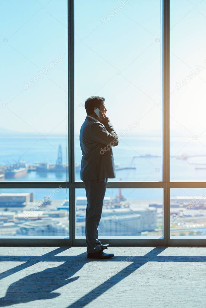 Mature business leader standing and looking out of large windows in a top floor office, talking on his mobile phone while looking out at the view