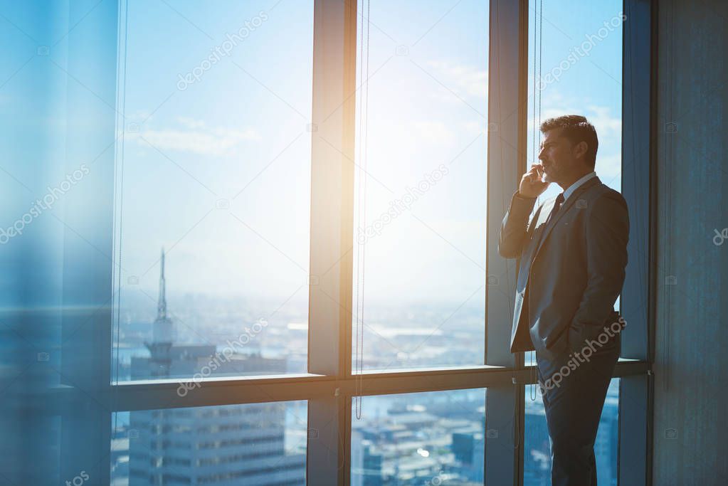 Handsome mature businessman standing in a professional corporate suit alongside large windows with a view of the city, talking on his mobile phone and looking out 