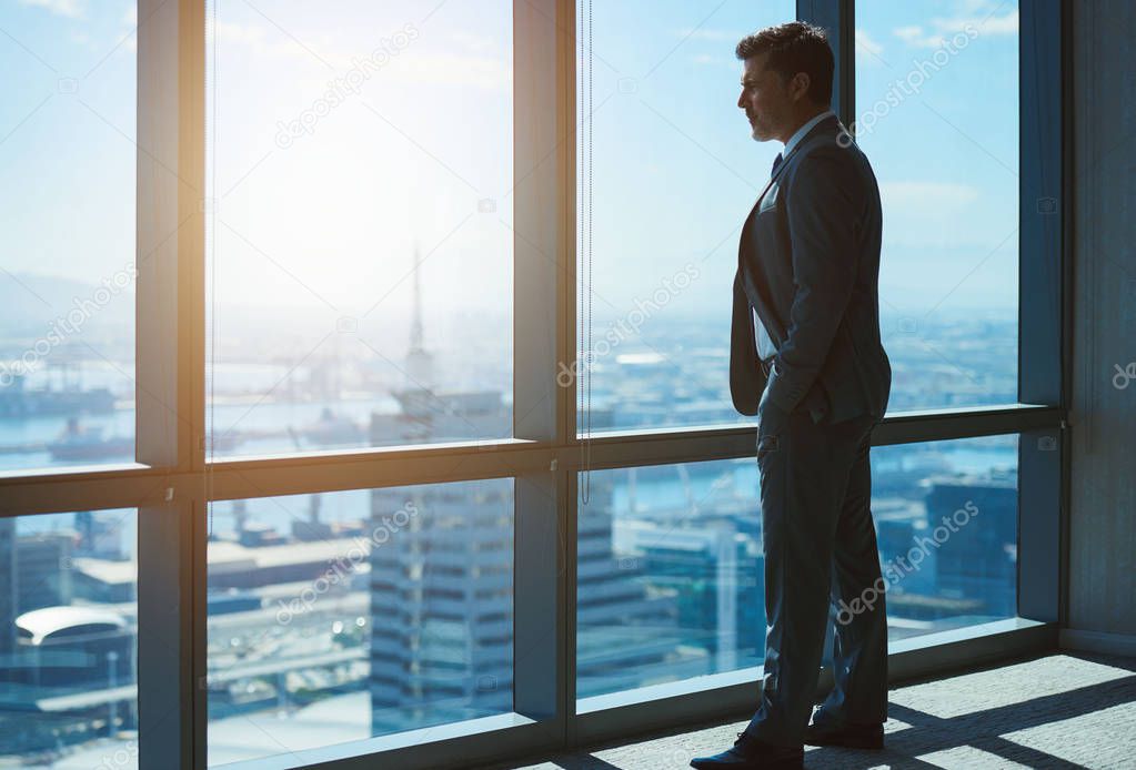 Mature business executive and leader standing confidently with hands in his pockets, in the the top floor office, looking at the city below through large windows