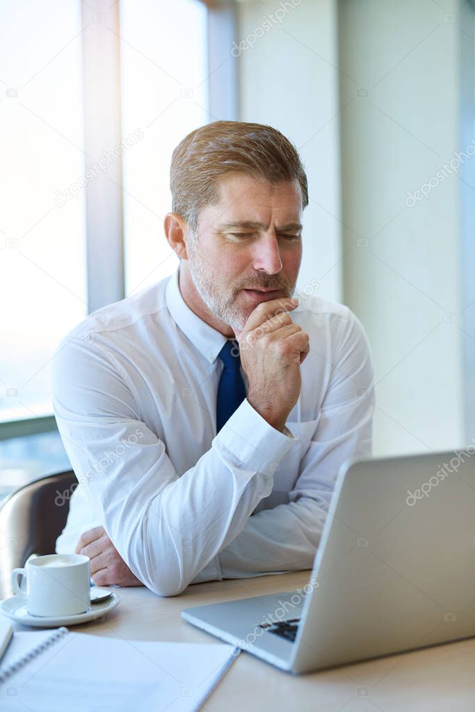 Handsome and mature businessman sitting at a desk with a cup of coffee and holding his chin, while looking seriously at information on the screen of his laptop computer