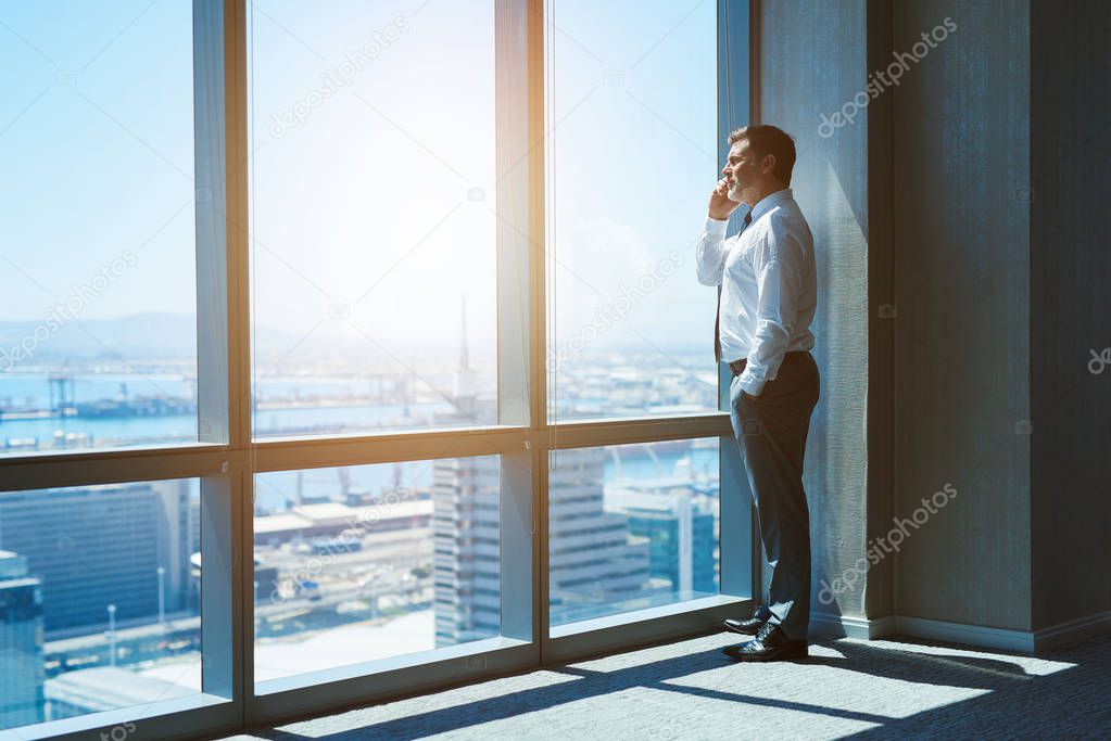 Mature and cnofident business executive looking looking out of large windows at a view of the city below, from the top floor of an office building, while talking on his mobile phone