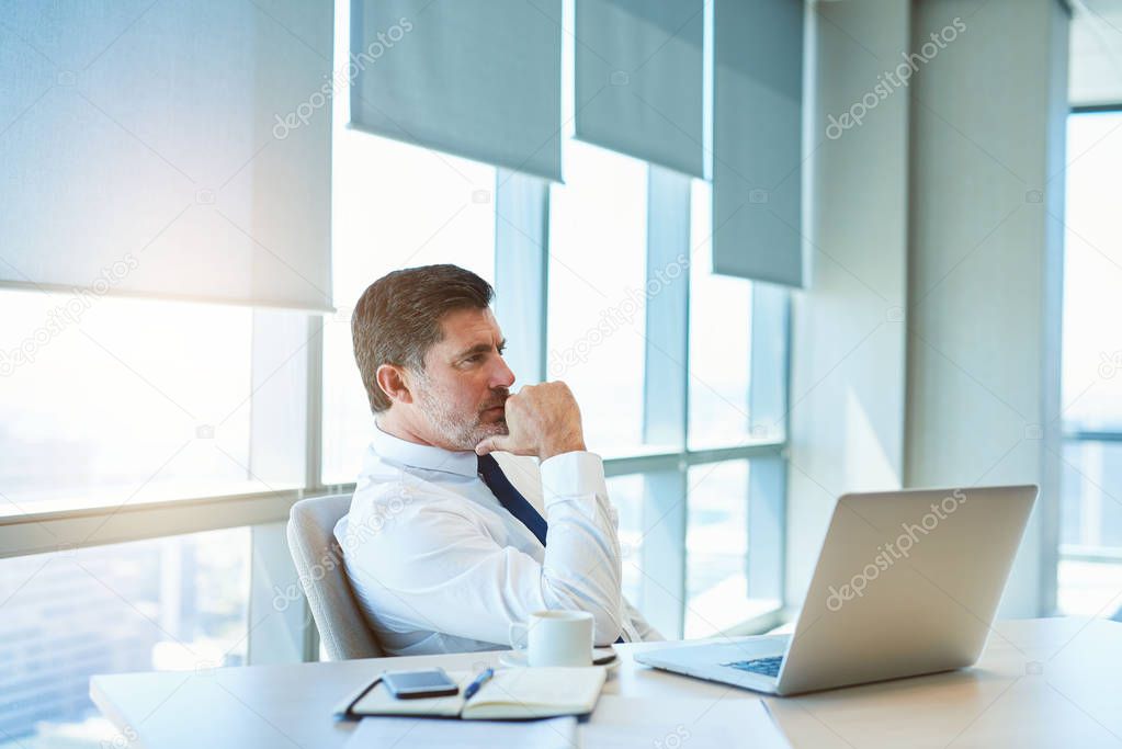 Handsome mature business manager sitting at his desk in a modern office with his laptop open in front of him looking away with a deeply thoughtful expression.