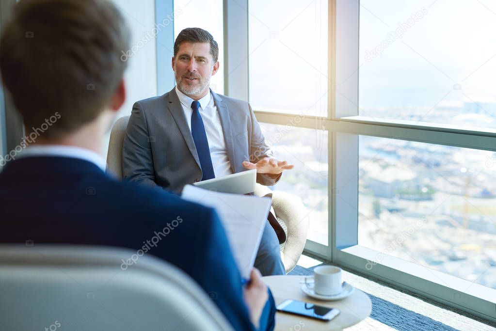 Handome mature corporate manager sitting in a modern office space, holding a digital tablet while interviewing a young business applicant