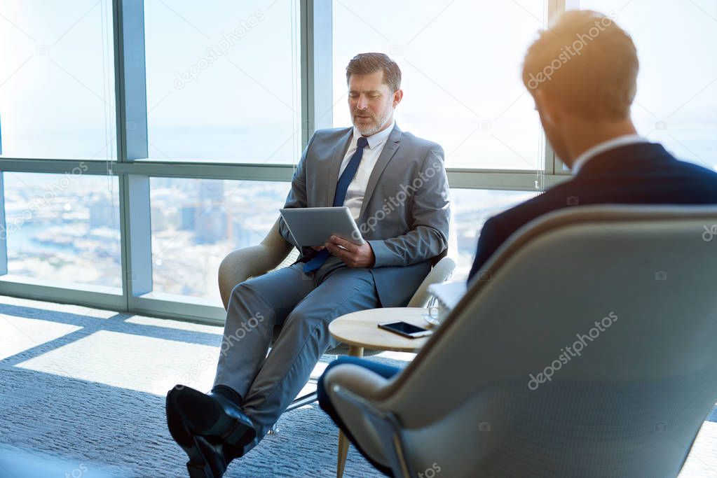 Handsome mature businessman sitting in a modern office space, using a digital tablet, with a younger man making notes 