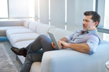 Mature businessman sitting in a relaxed position on a modern couch, using a digital tablet with a positive expression