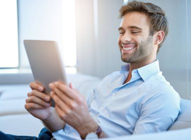 Handsome young man wearing a business shirt, sitting on a couch and laughing while reading something entertaining on his digital tablet