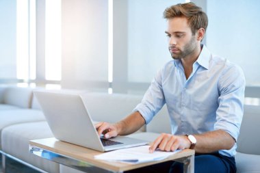 Handsome young businessman with designer stubble, sitting in a modern business lounge working on his laptop computer