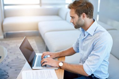 Genç ve yakışıklı bir iş adamının modern bir iş salonunda güneşlenirken dizüstü bilgisayarı üzerinde çalışmasının yüksek açılı görüntüsü.