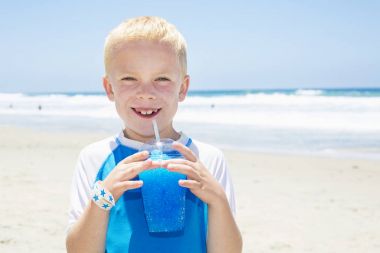 Young boy drinking a blue ice drink at the beach during summer vacation clipart