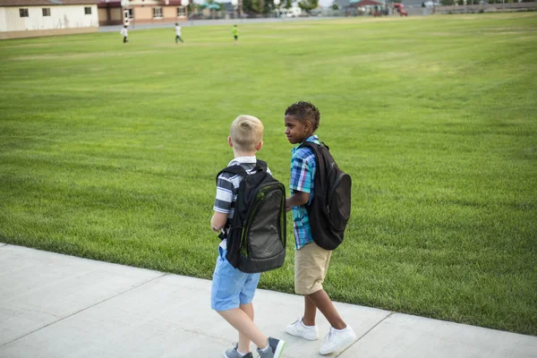 2人の多様な学校の子供たちが歩いて学校に行く途中で話し合っています バックパックを着た多様な学校の子供たちの学校の写真に戻る — ストック写真