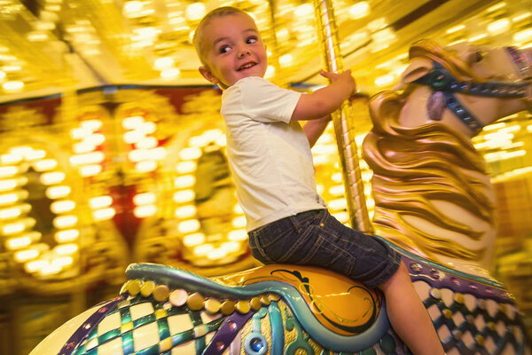 Cute little boy having fun riding a carousel at an amusement park or carnival. Happy little boy riding a merry go round carousel with bright lights