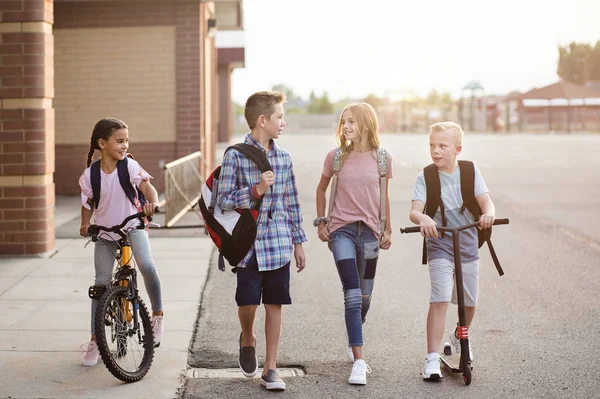 学校の子供たちの多様なグループが話し 一緒に学校から家に歩いています 男の子と女の子の両方の完全な長さの率直な写真彼らの自転車やスクーターで学校の建物を離れる — ストック写真