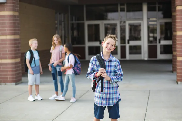 学校の後に友達とぶらついているハンサムな未成年の少年学生 学校の建物の外に立っている笑顔の男の子に焦点を当て 背景に友人と — ストック写真