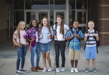Okul binasının önünde gülümseyen ergenlik öncesi öğrencilerin geniş bir grup portresi. Sırt çantaları takan ve okula gitmeye hazır bir grup çocuğun okul fotoğrafı.