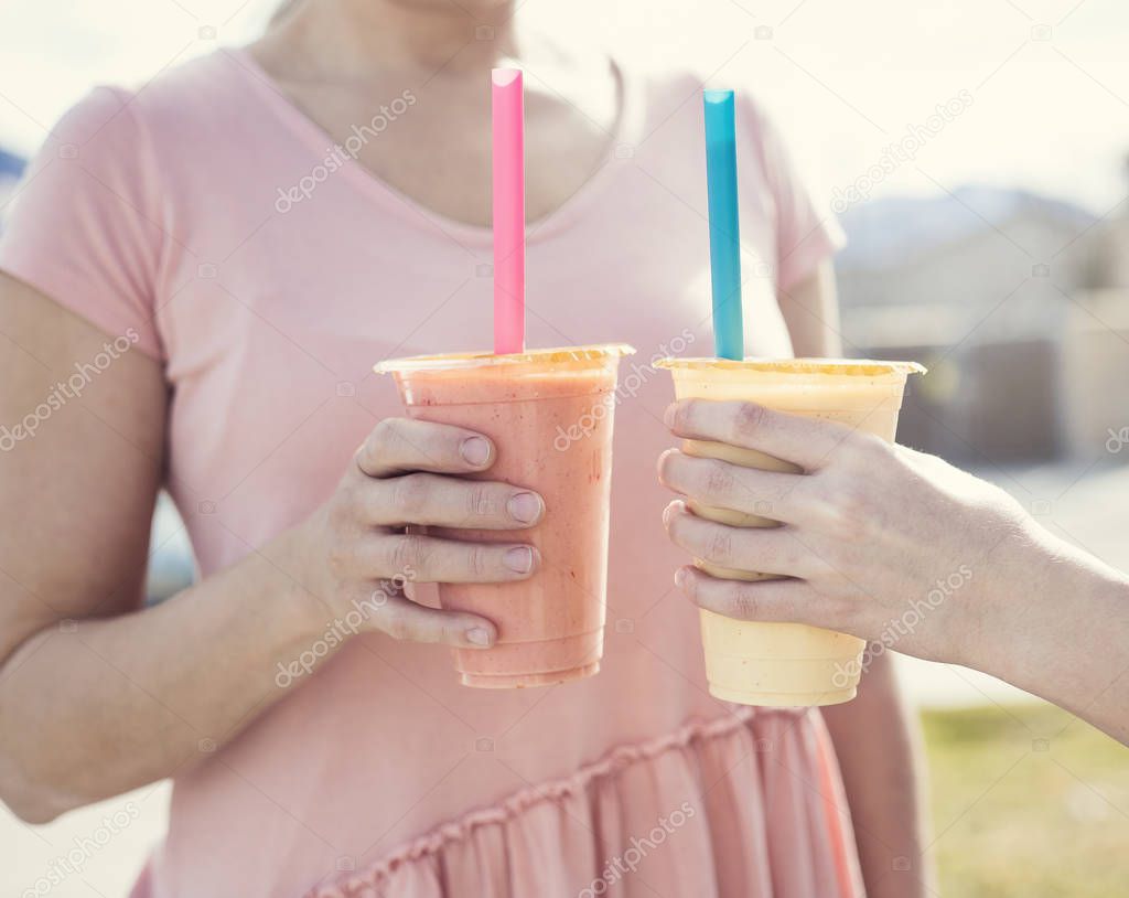 Closeup of two people holding healthy fruit smoothie drinks in their hands. Outdoor photos with focus on the smoothies. Eating healthy and treating your body well