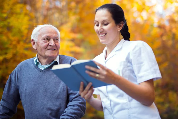 Enfermera sonriente leyendo libro a hombre mayor que utiliza andador con c — Foto de Stock