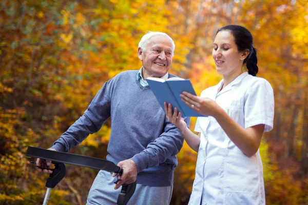 Enfermera sonriente leyendo libro a hombre mayor que utiliza andador con c — Foto de Stock